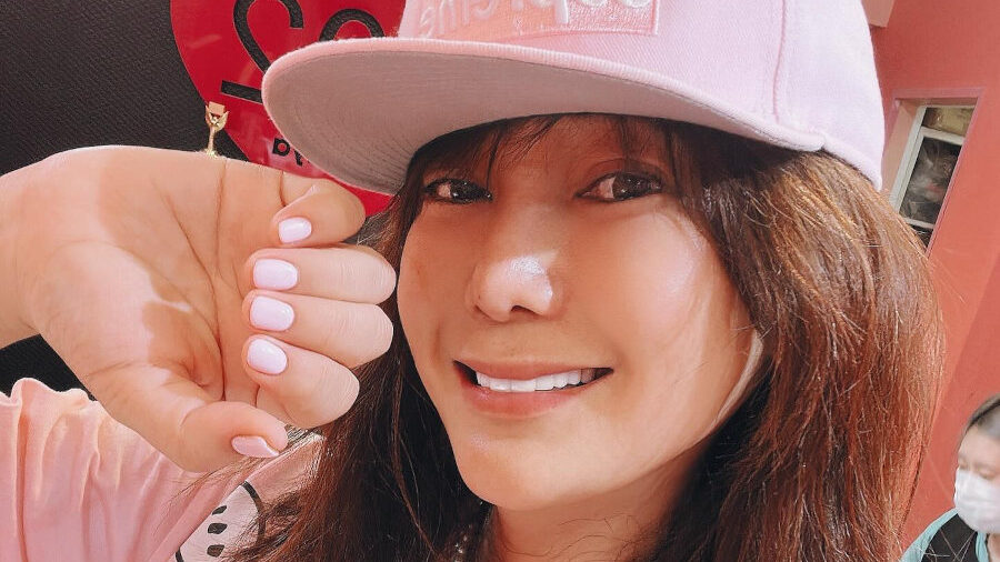 Ai Haruna souriante avec un t-shirt et une casquette roses, montrant ses ongles vernis.